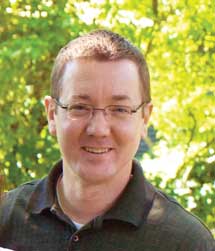 Photo of Ken Floody, a bespectacled man, standing in front of trees in the sunlight