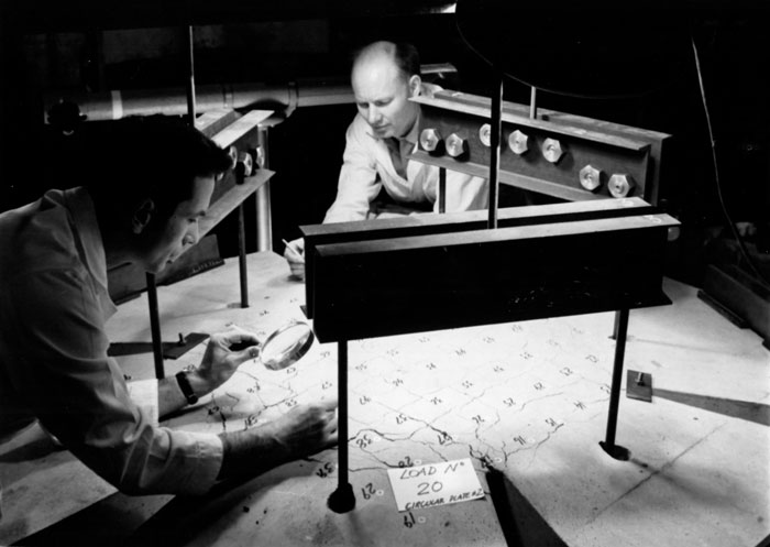 Photo from the archives: A circular plate with wings, made of reinforced concrete is subjected to bending and twisting moments in more than one direction. The deflections are measured by a combination of photogrammetry and mechanical dial gages by Prof. M. A. Sozen (left) and Mr. R. J. Lenschow.